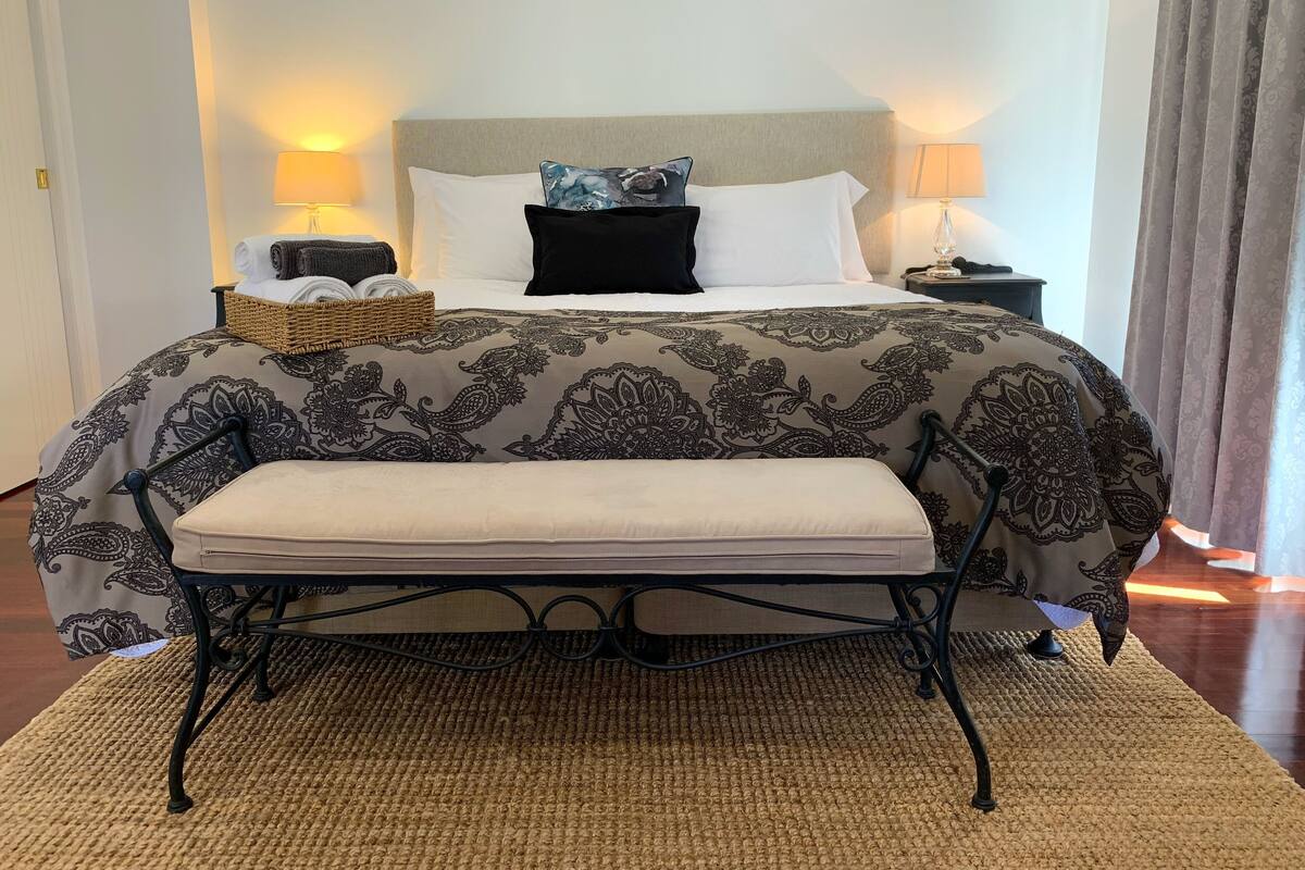 A neatly made bed with a patterned bedspread, a bedside table with a lamp, and a bench at the foot of the bed.