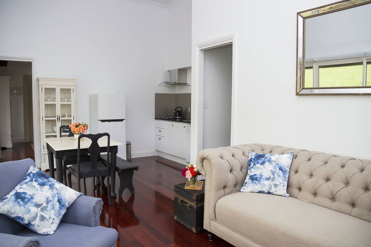 A living room with a beige sofa, two blue floral cushions, a small table with a vase of flowers, and a dining area with a white cabinet and black chairs.