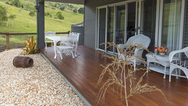 A covered outdoor patio with a wooden deck, white wicker furniture, and a view of a green hillside. - Country Mile Escape (Flying Fox)