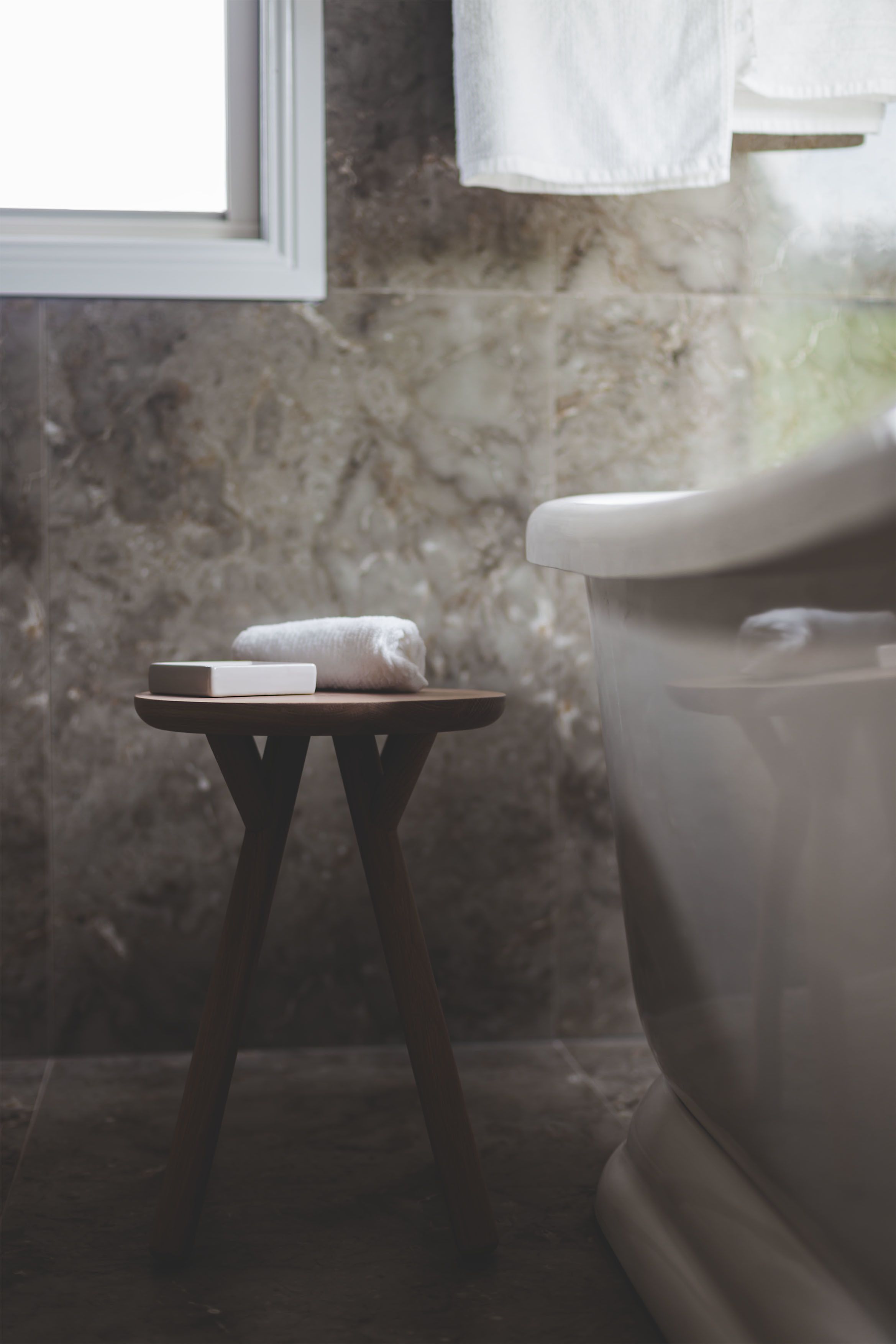 A bathroom with a marble wall, a white toilet, a small wooden stool with a white towel, and a framed picture above the toilet.