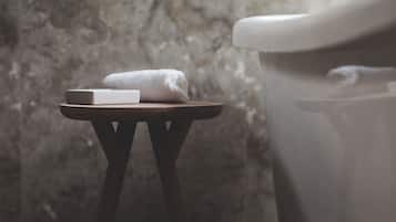 A bathroom with a marble wall, a white toilet, a small wooden stool with a white towel, and a framed picture above the toilet.