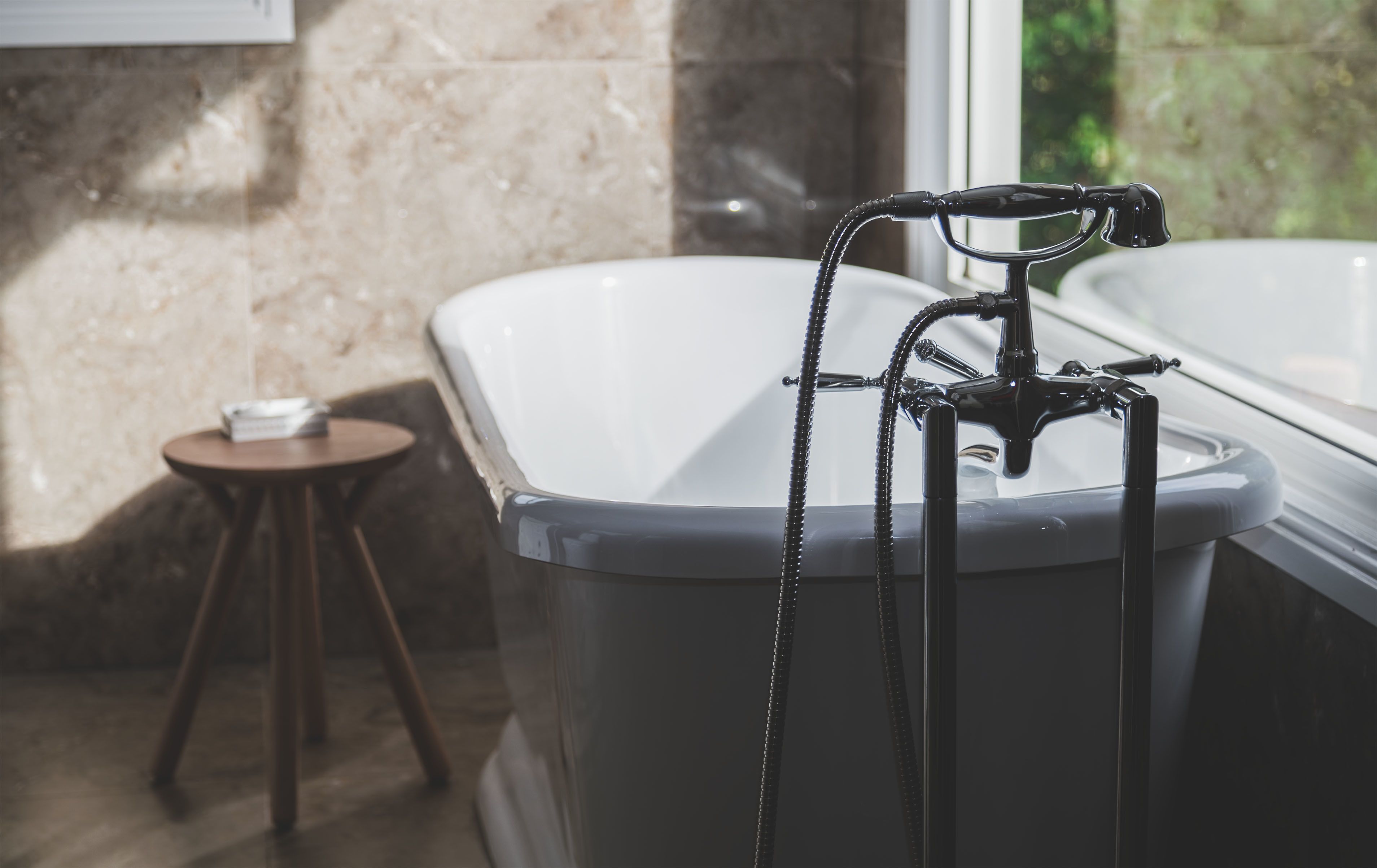 A modern bathroom with a freestanding bathtub, a small round table with a cup, and a view of greenery outside.