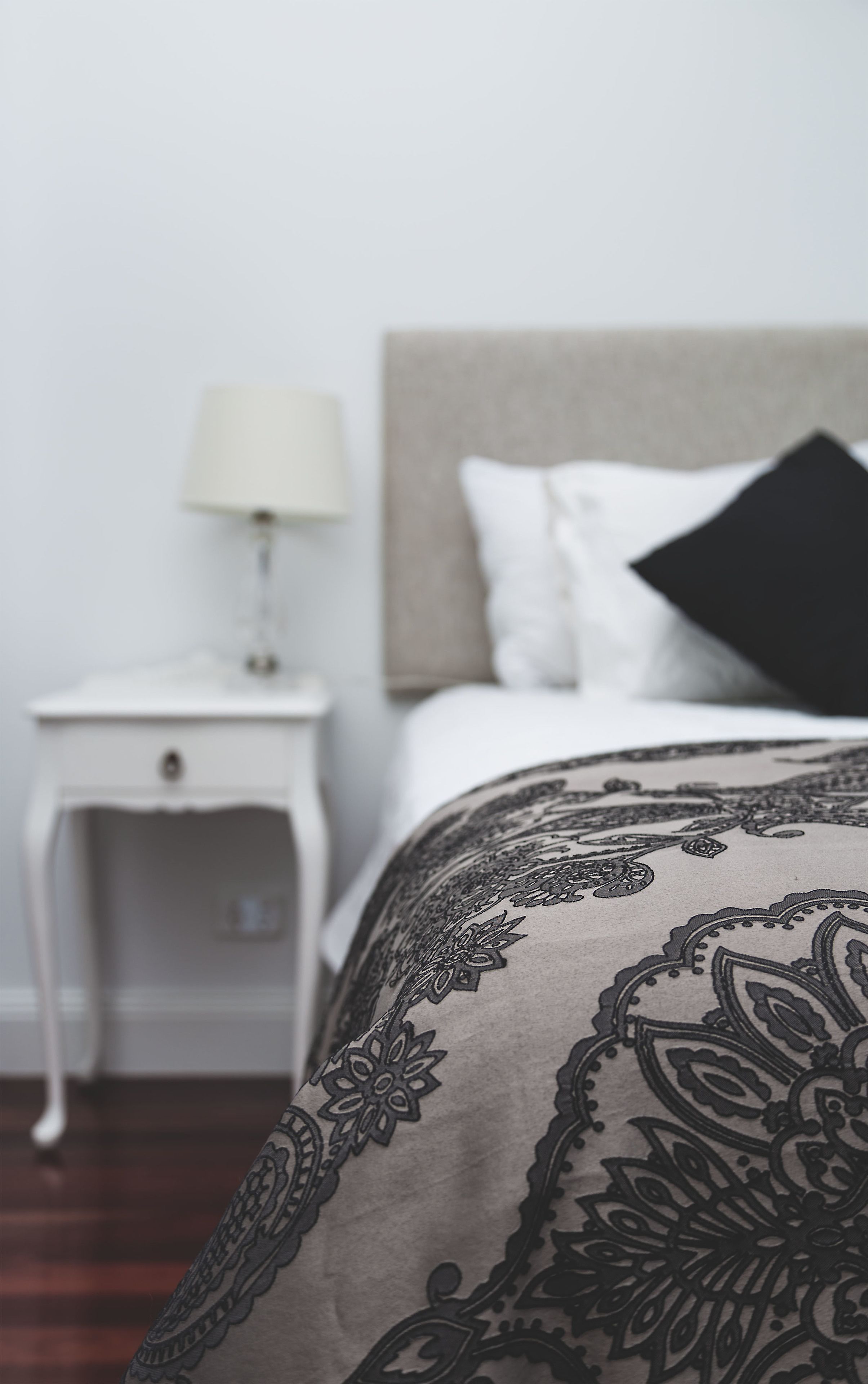 A neatly made bed with a patterned bedspread, a white nightstand with a lamp, and a headboard with pillows.