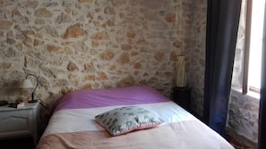 A room with a stone wall, a bed with a purple and white bedspread, a patterned pillow, a nightstand with a lamp, and a window with curtains. - In the heart of Provence, near the verdon stone house of character (Varages)
