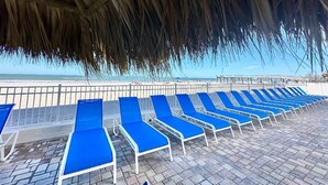 Un área de descanso frente a la playa con sillones azules bajo un techo de paja, con vista al mar.