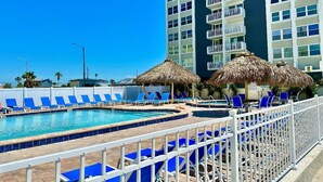 Zona de piscina con sillas de descanso azules y sombrillas de paja, rodeada por una valla blanca.