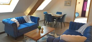 Ein gemütlicher Loftsraum mit schrägem Dach, einem Essbereich mit blauen Stühlen und einem blauen Sofa mit weißer Decke.