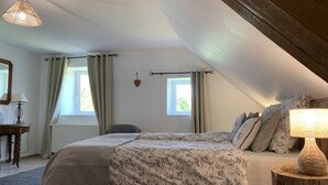 Un dormitorio con techo inclinado, cama con ropa de cama floral, lámpara de noche, mesita de noche de madera y ventana con cortinas.
