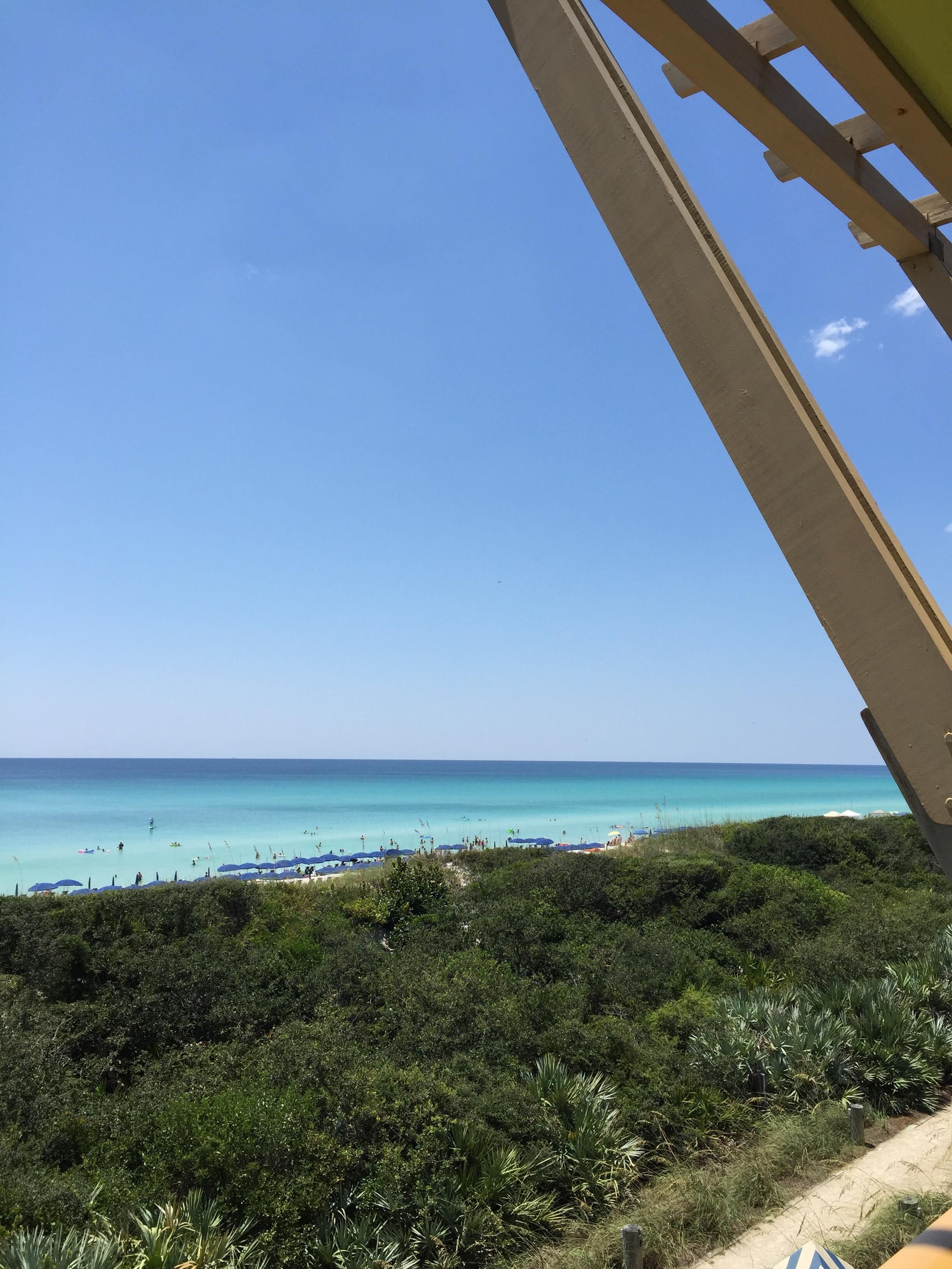 A beach with clear blue water, umbrellas, and dense greenery in the foreground.