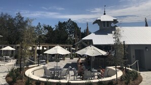 An outdoor patio area with round tables and chairs, umbrellas, and a building with a distinctive tower. - Steps away from Camp Watercolor Amenities  (Santa Rosa Beach)