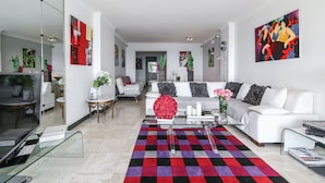 Una sala de estar moderna con un sofá blanco, una mesa de centro de vidrio y una alfombra colorida. Hay cuadros en las paredes y un espejo grande que refleja la habitación.