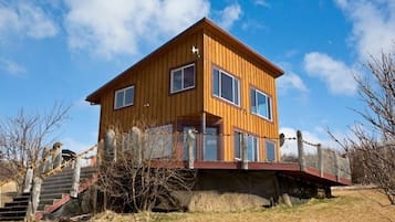 Une maison en bois à deux étages, dotée d’un balcon, entourée d’une zone délimitée par une clôture et de quelques plantes.