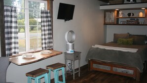 A cozy room with a bed, a small table with a mirror, a TV mounted on the wall, and a window with curtains. - Tiny Red Farmhouse centrally located in Columbia Falls (Columbia Falls)