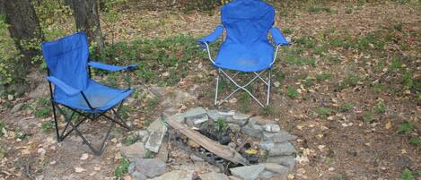 Two blue folding chairs and a stone fire pit in a wooded area.
