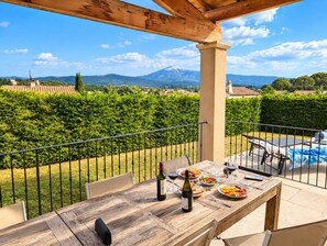 Une table en bois avec des sièges pour l’extérieur, une piscine et un paysage montagneux en arrière-plan.