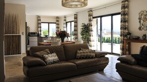 A modern living room with a brown leather sofa, a dining area, and large windows offering a view of the city.