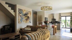 A modern living room with a sofa, coffee table, and a staircase. There is a dining area with a table and chairs, and a kitchen in the background.