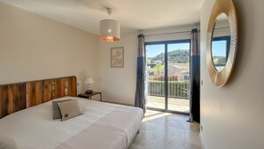 A bedroom with a bed, a nightstand, a lamp, a framed picture, a large mirror, and a balcony with a view of houses and trees.