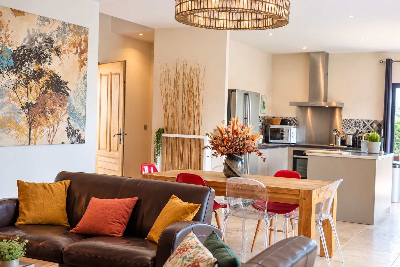 A modern living room with a brown leather sofa, a dining area with a wooden table and red chairs, and a kitchen with stainless steel appliances.