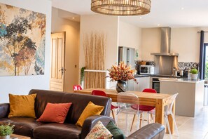 Una sala moderna con un sofá de cuero marrón, un comedor con una mesa de madera y sillas rojas, y una cocina con electrodomésticos de acero inoxidable.