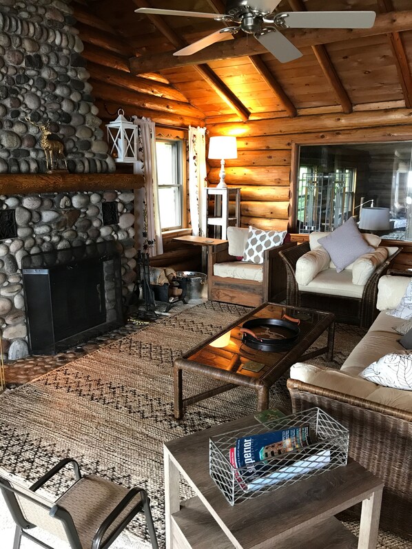 Een gezellig chaletinterieur met een stenen openhaard, houten wanden, een plafondventilator en een comfortabele zithoek met een bank en fauteuils.