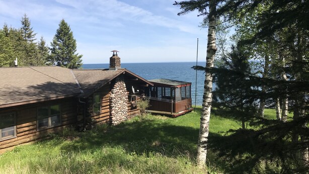 Une cabane en rondins avec une cheminée en pierre, une terrasse en bois et une vue sur l’eau.