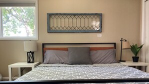 A neatly made bed with a patterned bedspread, a headboard with a geometric mirror, a bedside lamp, a small table with a lamp, and a potted plant.