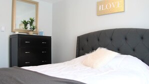 A bedroom with a tufted headboard, a bed with white bedding, a dark dresser, a mirror, and a '#LOVE' sign on the wall.