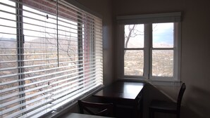 A corner room with a dining table and chairs, a window with blinds, and a view of a barren landscape.