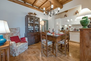 A dining area with a wooden table, chairs, and a chandelier. There is a cabinet with glass doors and a floral sofa with red cushions.