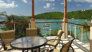 A balcony with a glass table and wicker chairs, overlooking a marina with boats and a green hillside.