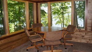Una cabaña de madera con una mesa redonda y cuatro sillas, ventanas amplias con vistas a árboles y agua.