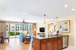 A modern hotel room with a large sliding glass door leading to a balcony, a kitchen area with a central island, and a seating area with a sofa and chairs.