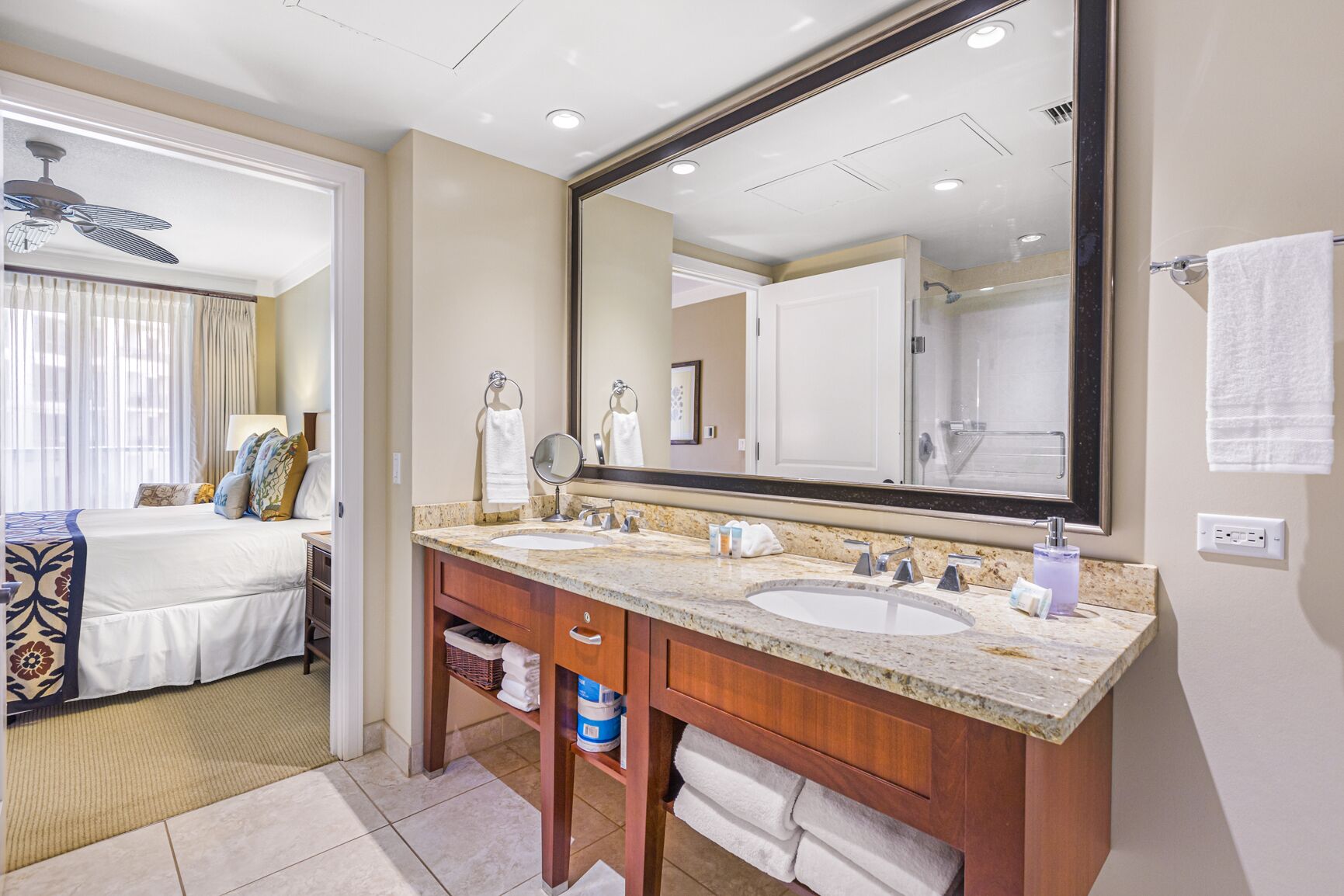 Une salle de bain d’hôtel avec deux lavabos, un grand miroir et un lit visible à travers une porte ouverte.