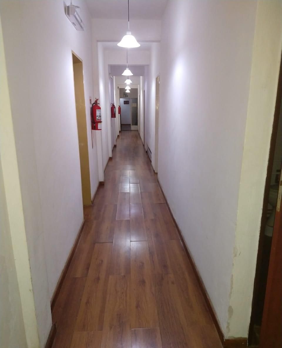 A long, narrow hallway with wooden flooring and white walls. There are ceiling lights and fire extinguishers visible.