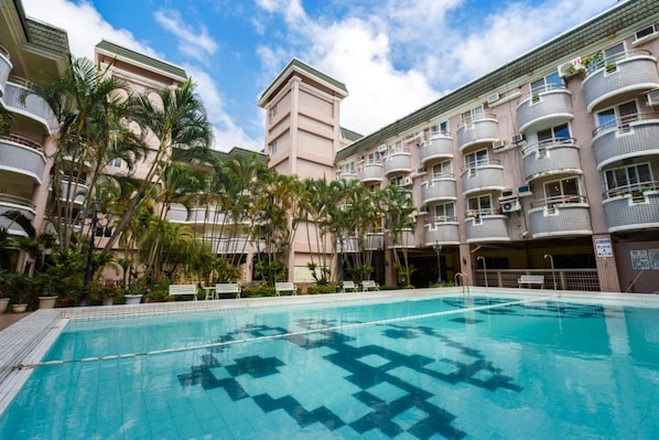 Una piscina rodeada de edificios de varios pisos con balcones, palmeras y un cielo despejado.