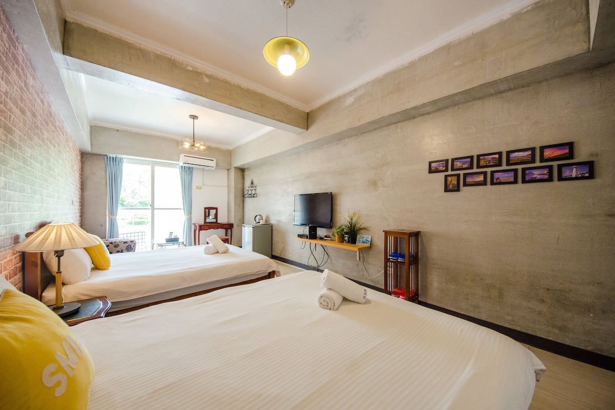 A hotel room with two beds, a TV, a shelf with pictures, and a window with curtains.