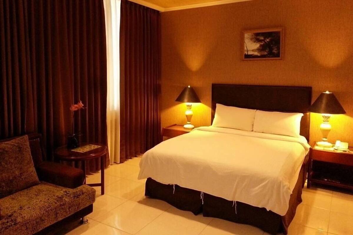 A hotel room with a bed, two bedside tables with lamps, a chair, a window with curtains, and a painting on the wall.