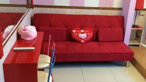 A red sofa with heart-shaped pillows, a small table with a remote control, and a white fan in a room with wooden flooring. - Landscape Rural B&B (Checheng)
