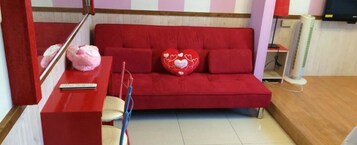 Un sofá rojo con almohadas con forma de corazón, una mesita con un control remoto y un ventilador blanco en una habitación con suelo de madera.