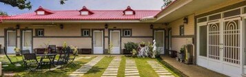 Un edificio de dos pisos con techo rojo, un porche cubierto con mesa y sillas, y un pequeño jardín.