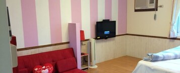 Una habitación con un sofá rojo, una mesa blanca con mantel azul, un televisor montado en la pared y una ventana con cortinas.