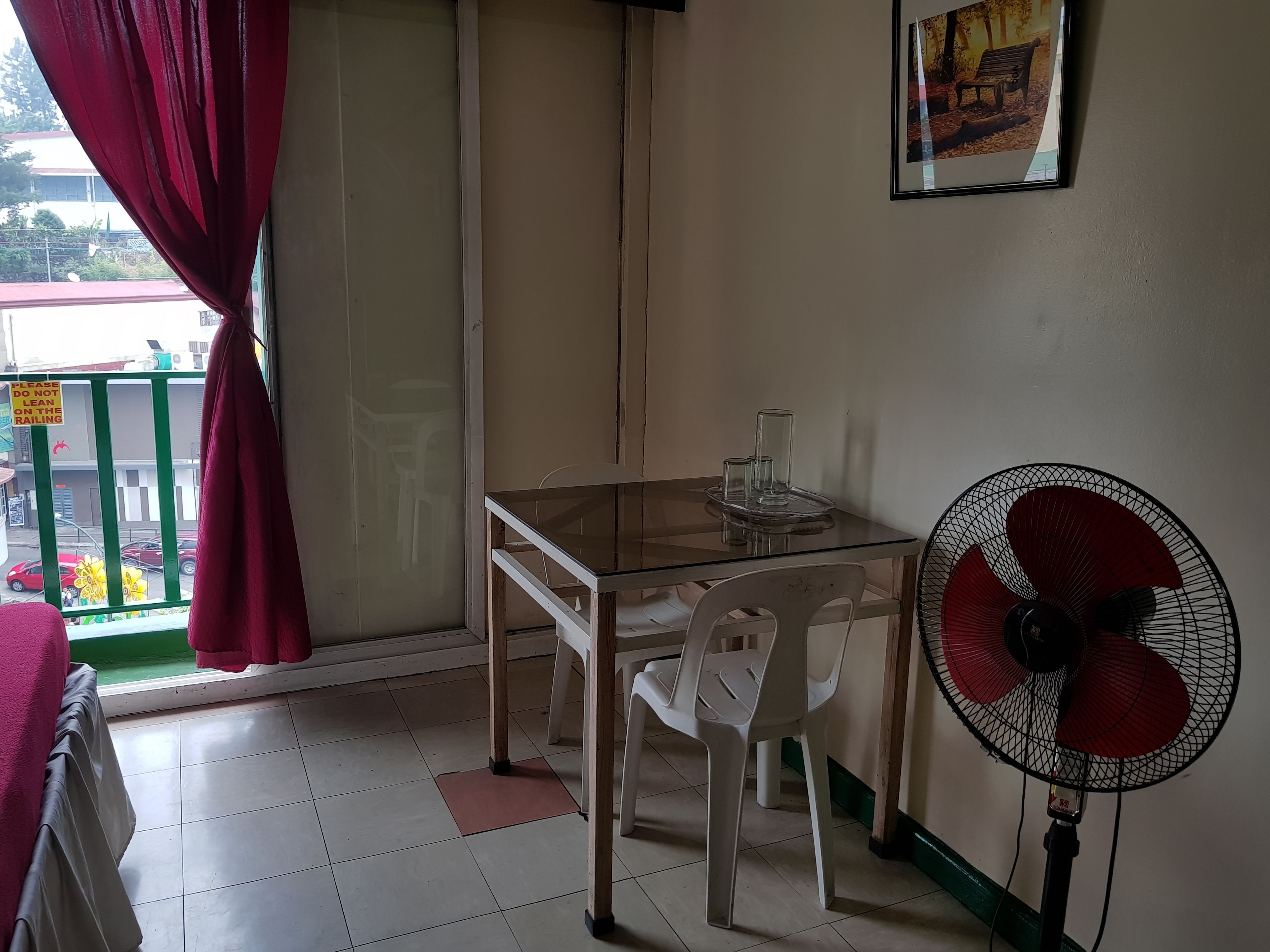 Una habitación pequeña con una mesa de vidrio, sillas blancas, una cortina roja, un ventilador de pared y un cuadro enmarcado.