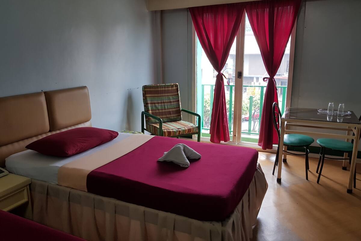 Une chambre d’hôtel avec un lit, une chaise, une table avec des verres et un balcon avec des rideaux rouges.