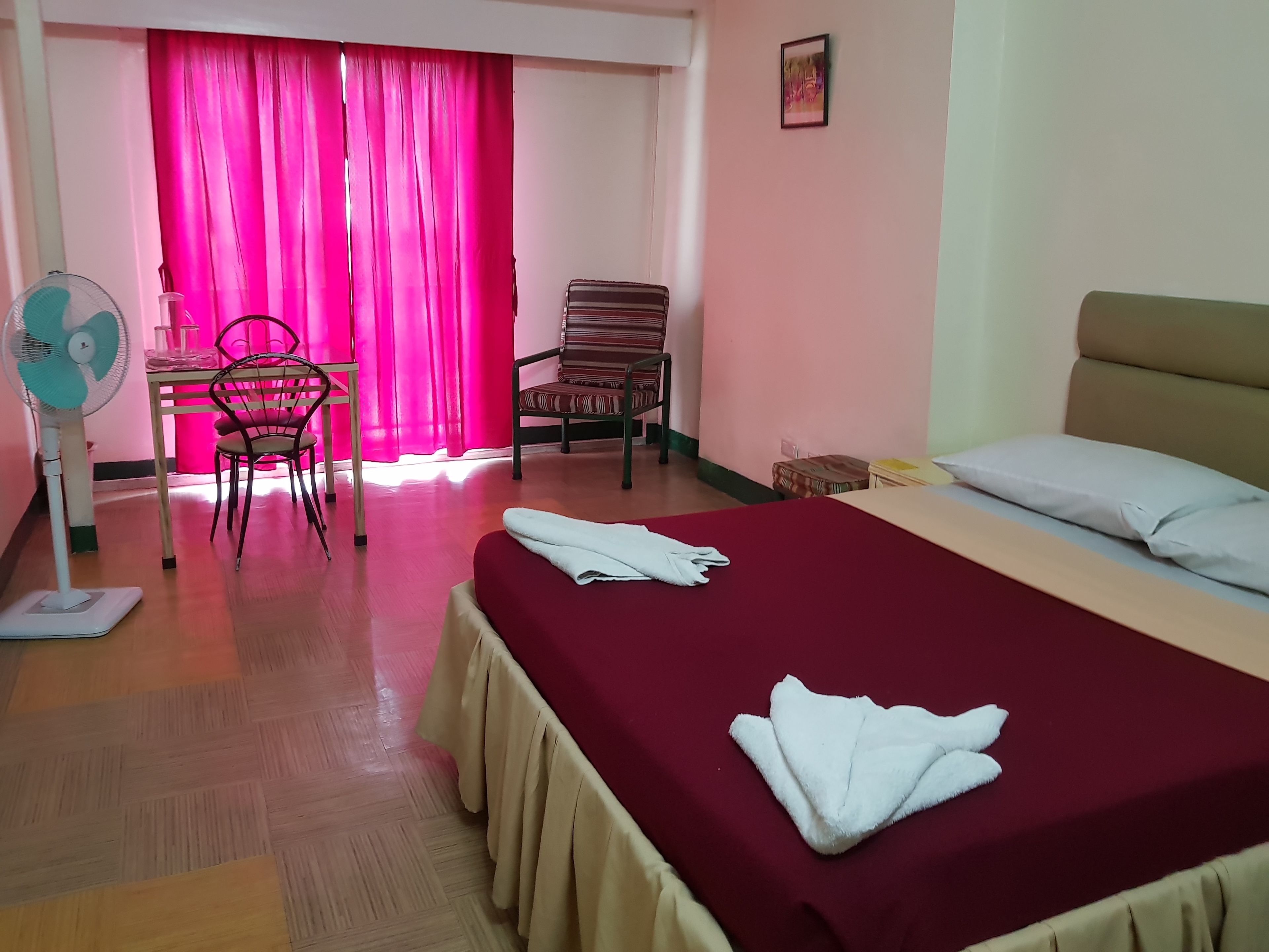 Habitación de hotel con cama, una silla, una mesa, un ventilador y una ventana con cortinas.