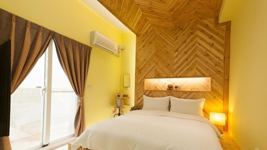 A hotel room with a bed, a nightstand, a wall-mounted air conditioner, a window with curtains, and a wooden ceiling.