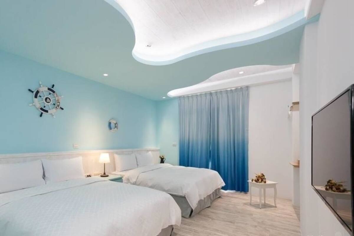A modern hotel room with two beds, a ship's wheel decoration, a flat-screen TV, and a large window with blue curtains.