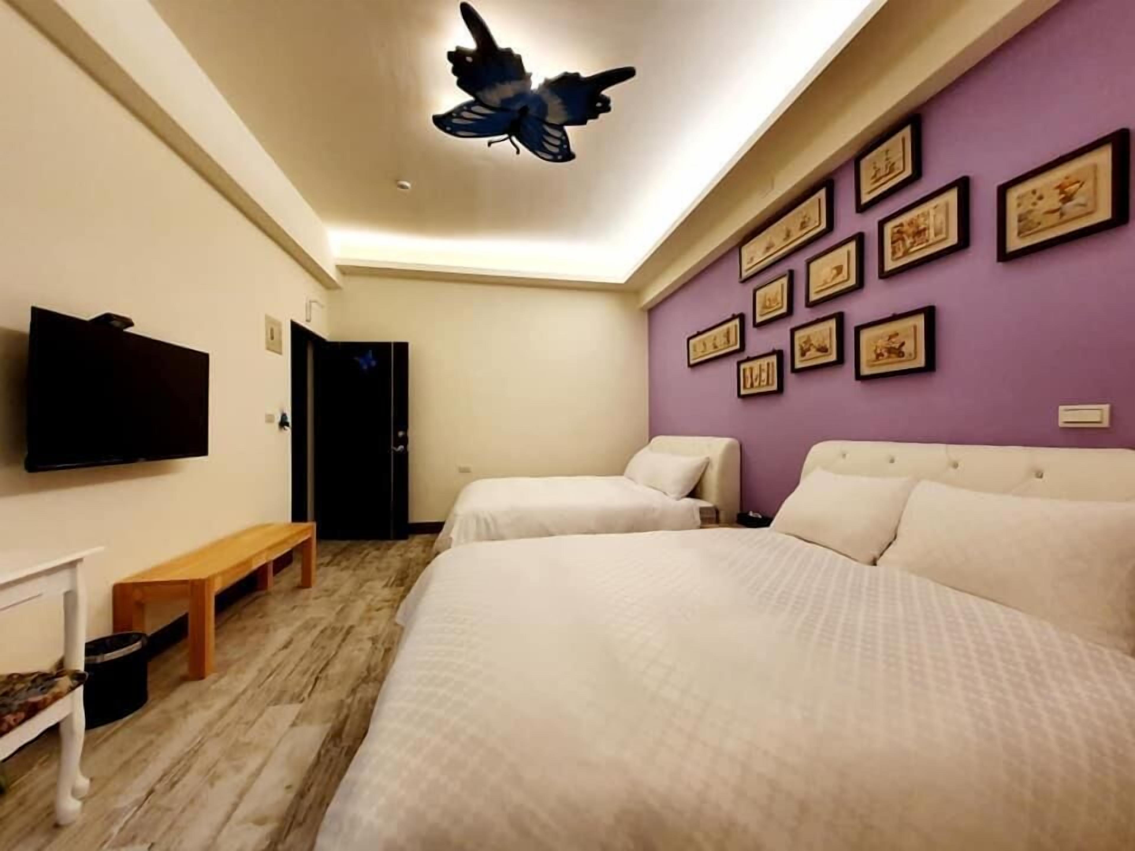 A hotel room with two beds, a flat-screen TV, a ceiling butterfly decoration, and framed pictures on the wall.