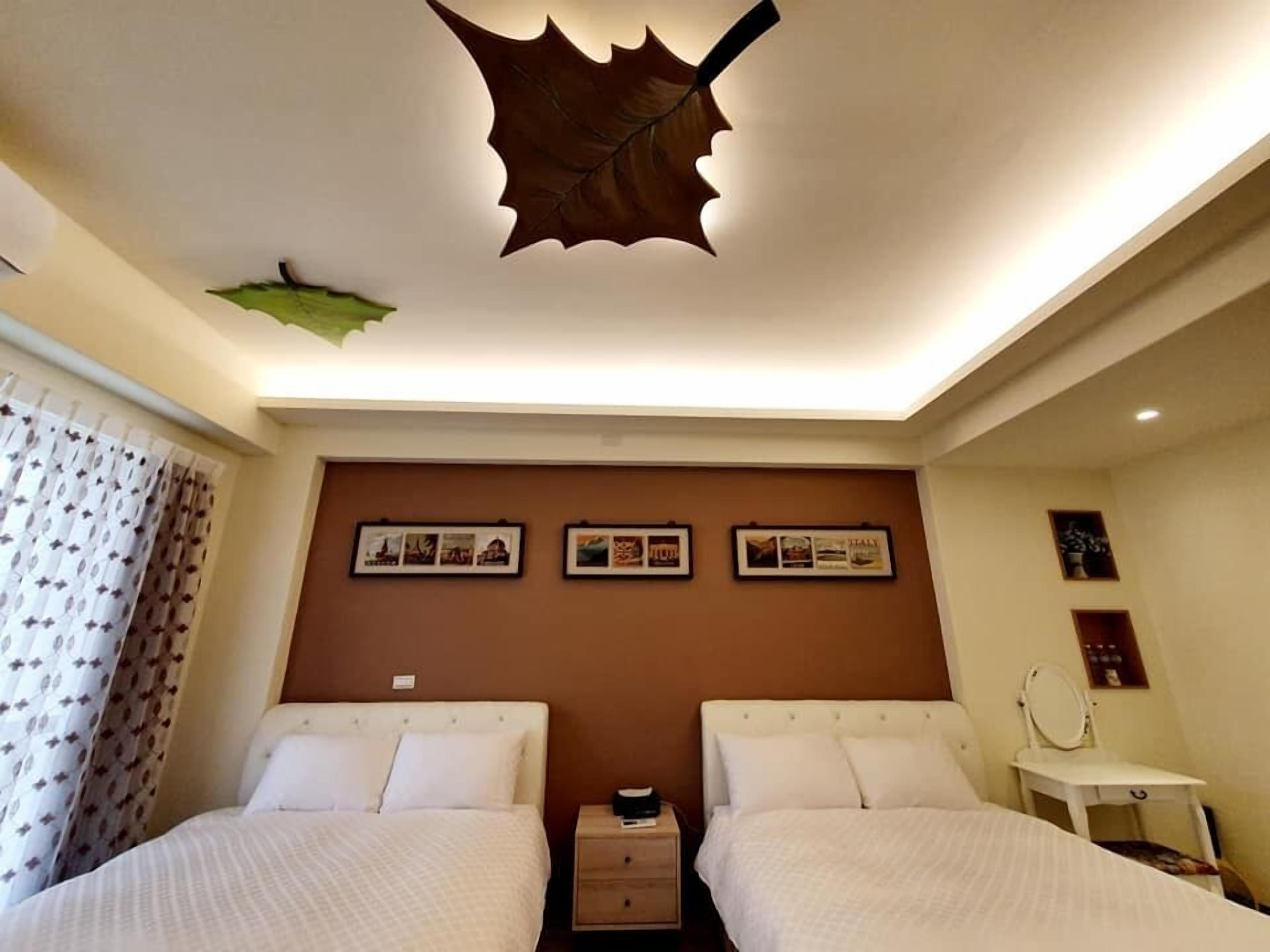 A hotel room with two beds, a leaf-shaped ceiling light, framed pictures on the wall, and a small bedside table with a mirror.