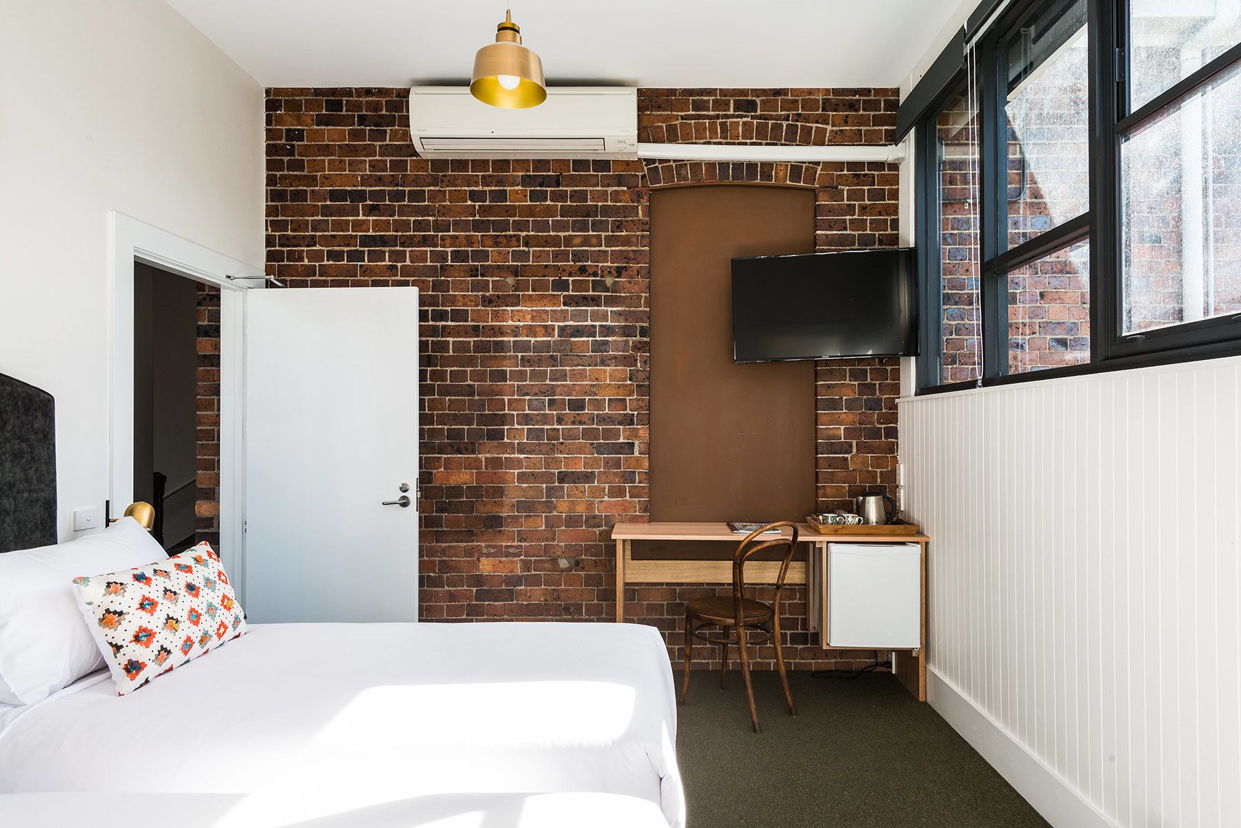 A hotel room with a brick wall, a bed with a white headboard, a desk with a chair, a television mounted on the wall, and a window with a view of the outside.